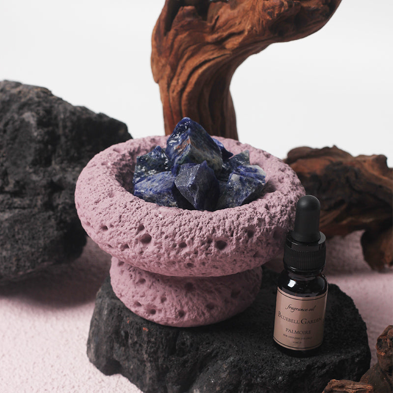 Close-up of purple stone Moon Vessel diffuser filled with blue crystals beside Bluebell Garden fragrance oil on dark natural rock surface