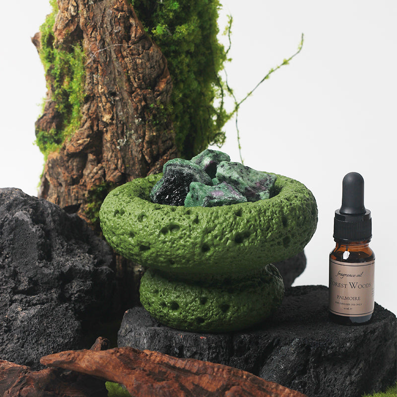 Green Moon Vessel diffuser with crystals styled with wood, stone, and greenery beside Forest Woods fragrance oil