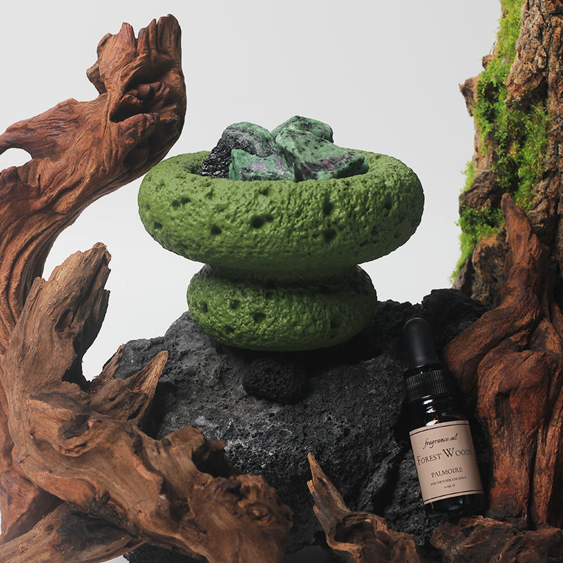 Green Moon Vessel diffuser with crystals styled with wood, stone, and greenery beside Forest Woods fragrance oil